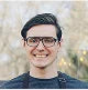 Person with glasses smiling outdoors; blurred background.